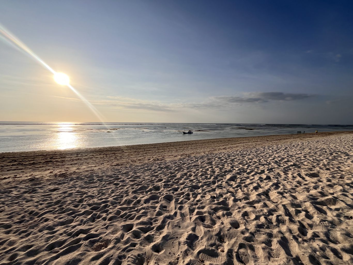 A wide, pristine white sand beach stretches towards calm, reflective turquoise-green waters under a bright sun with a lens flare. A small boat floats in the distance, and faint figures walk along the shore.