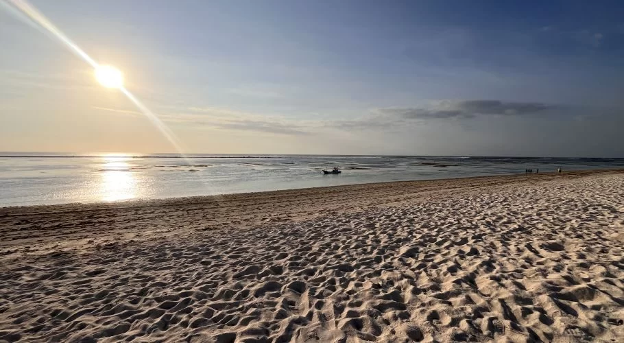 A wide, pristine white sand beach stretches towards calm, reflective turquoise-green waters under a bright sun with a lens flare. A small boat floats in the distance, and faint figures walk along the shore.