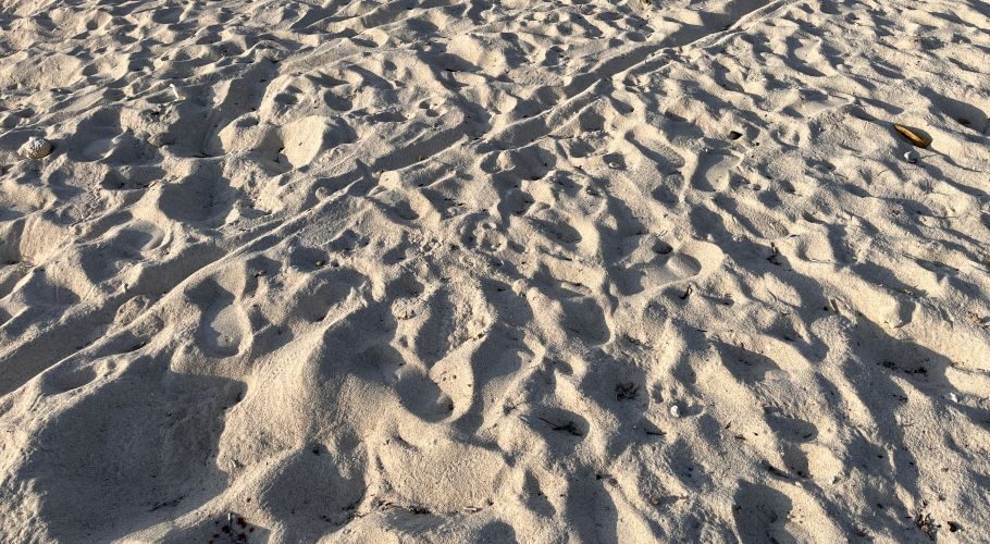 Pristine white sand on Tanjung Karoso Beach, covered in numerous footprints and ripples, casting long shadows under bright sunlight.