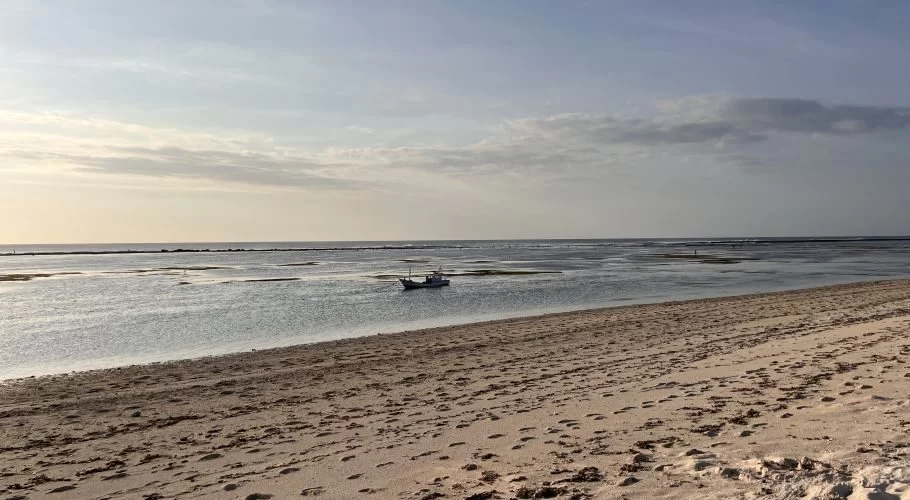 A pristine white sand beach meets clear turquoise-green shallow waters where a small fishing boat floats under a partly cloudy sky.