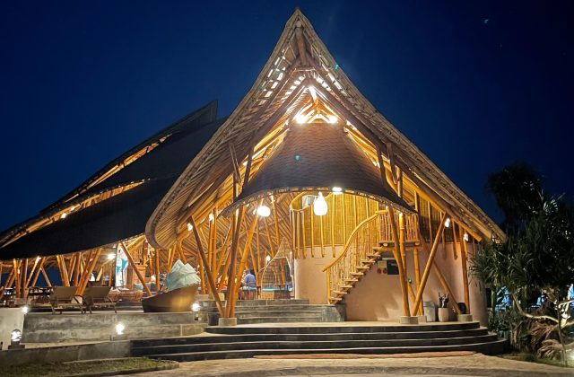 A large, multi-level bamboo resort building with peaked roofs and warm interior lighting stands illuminated against a dark blue night sky.