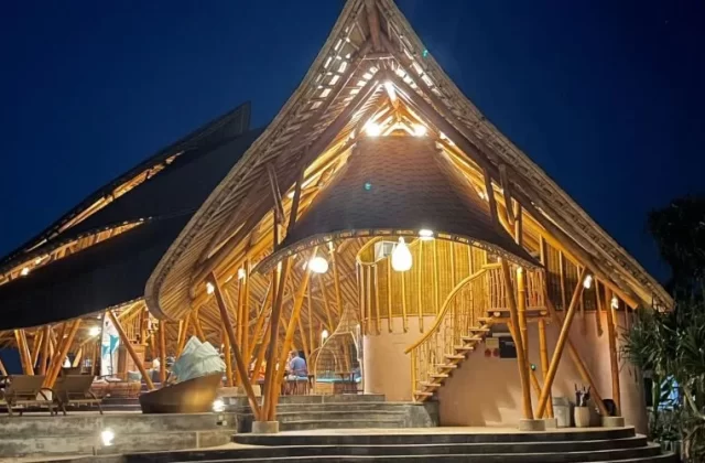 A large, multi-level bamboo resort building with peaked roofs and warm interior lighting stands illuminated against a dark blue night sky.