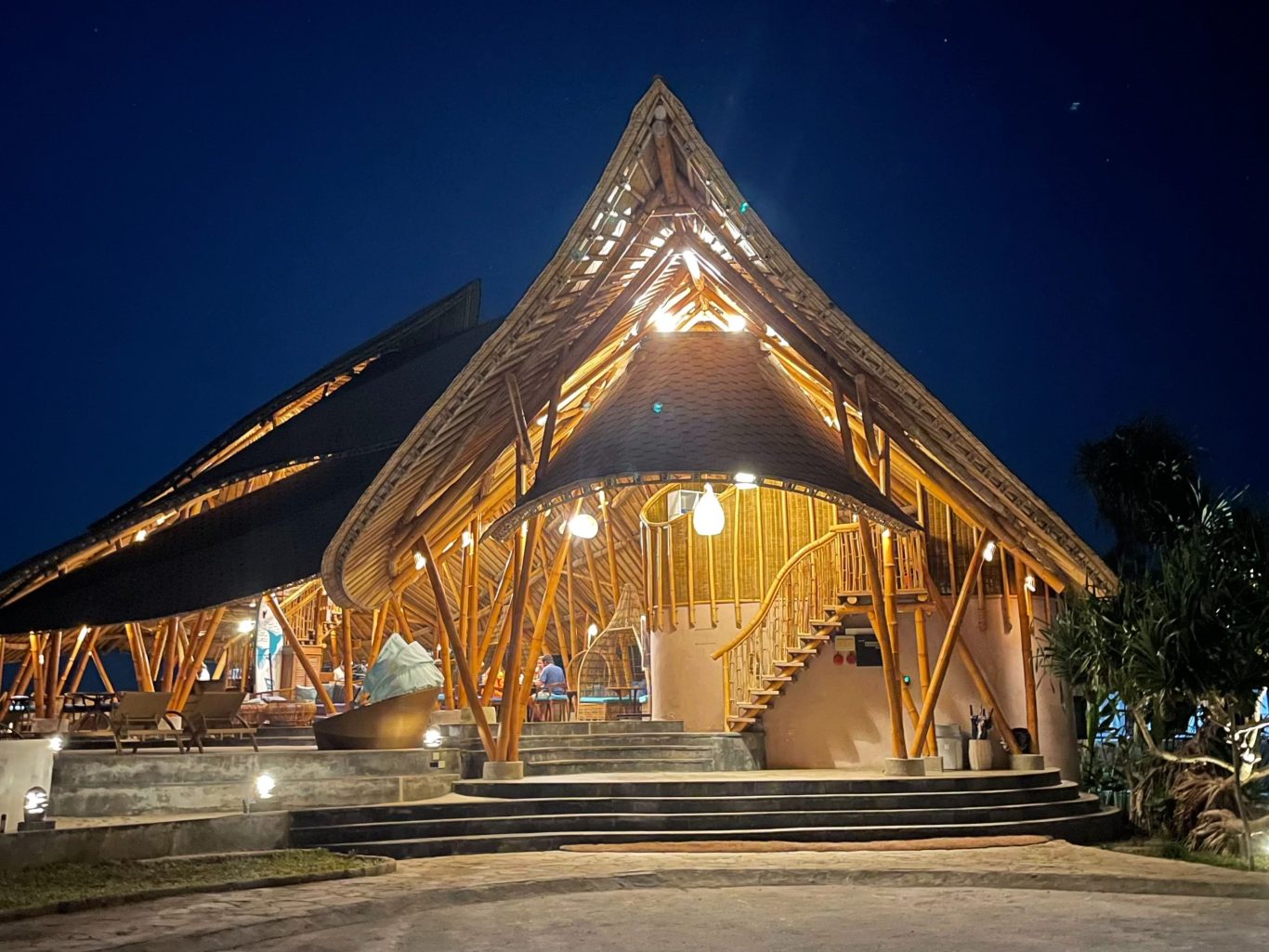 A large, multi-level bamboo resort building with peaked roofs and warm interior lighting stands illuminated against a dark blue night sky.