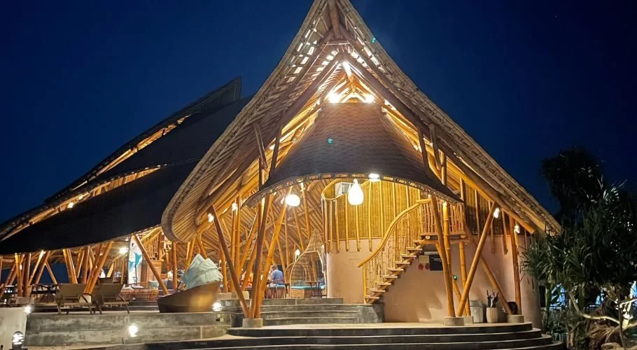 A large, multi-level bamboo resort building with peaked roofs and warm interior lighting stands illuminated against a dark blue night sky.