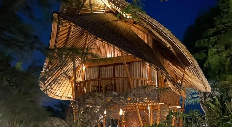 A two-story bamboo cottage with a curved thatched roof and a balcony is brightly lit from within at night, surrounded by dark tropical foliage.