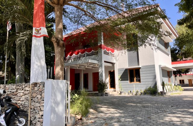 The modern, minimalist Tanto Hotel building in Kambera features white and gray walls with red accents, a paved parking area, and an Indonesian flag flying on the left.