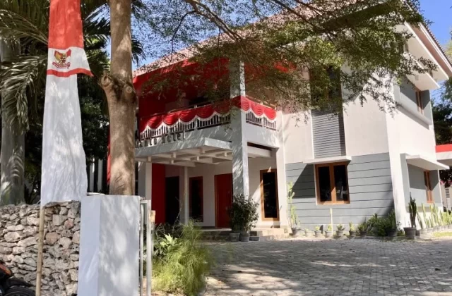 The modern, minimalist Tanto Hotel building in Kambera features white and gray walls with red accents, a paved parking area, and an Indonesian flag flying on the left.
