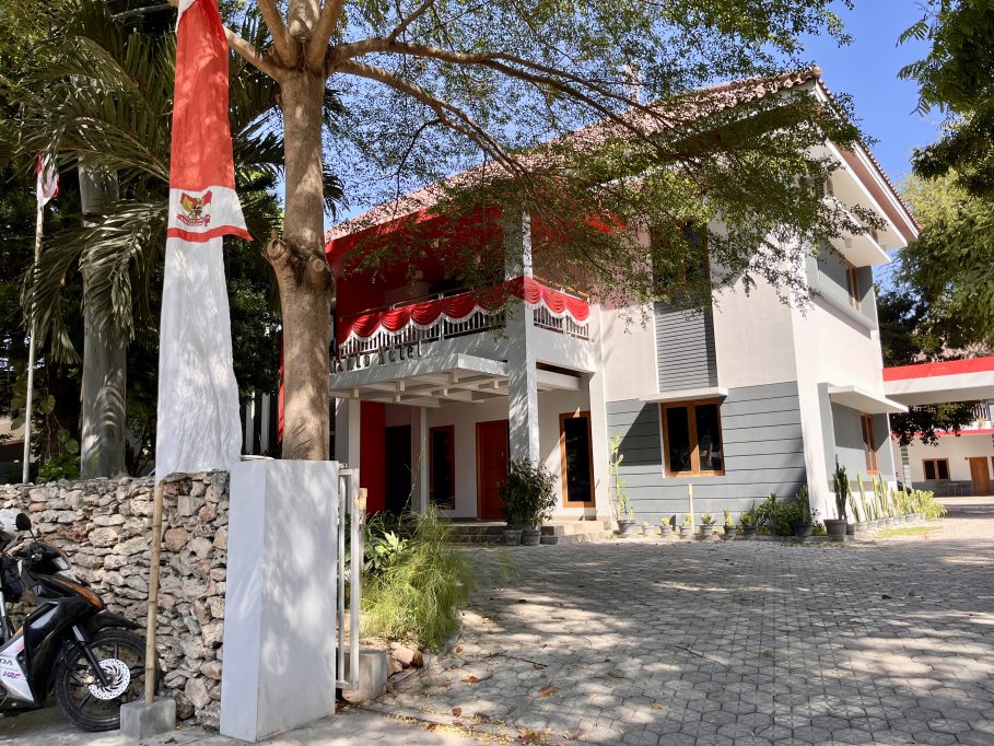 The modern, minimalist Tanto Hotel building in Kambera features white and gray walls with red accents, a paved parking area, and an Indonesian flag flying on the left.
