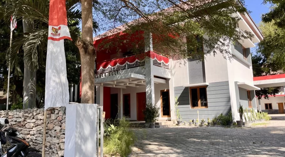 The modern, minimalist Tanto Hotel building in Kambera features white and gray walls with red accents, a paved parking area, and an Indonesian flag flying on the left.