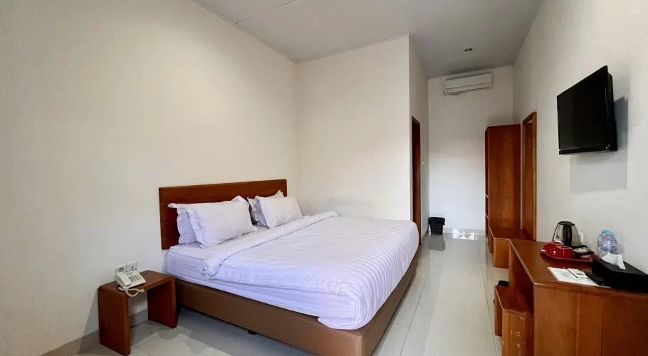 A modern hotel room features a bed with white linens, a wooden headboard, a wall-mounted TV, a desk, and a wardrobe, all against white walls and tiled floor.