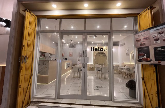 The brightly lit interior of a modern Es Teh Indonesia shop in Waingapu, Sumba, is visible through its glass doors, flanked by partially open yellow security gates.