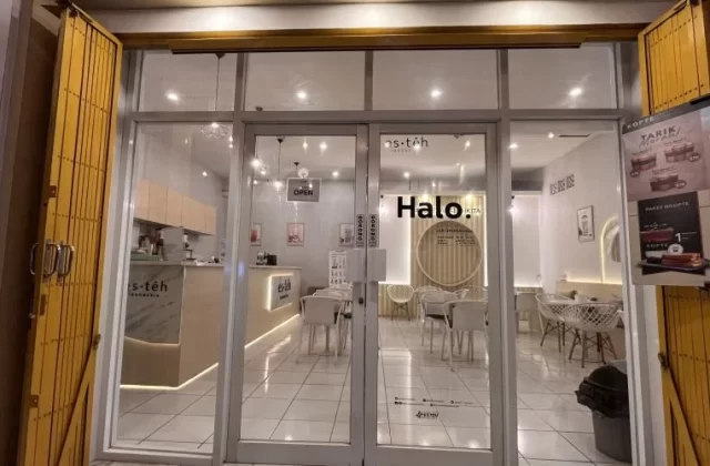 The brightly lit interior of a modern Es Teh Indonesia shop in Waingapu, Sumba, is visible through its glass doors, flanked by partially open yellow security gates.