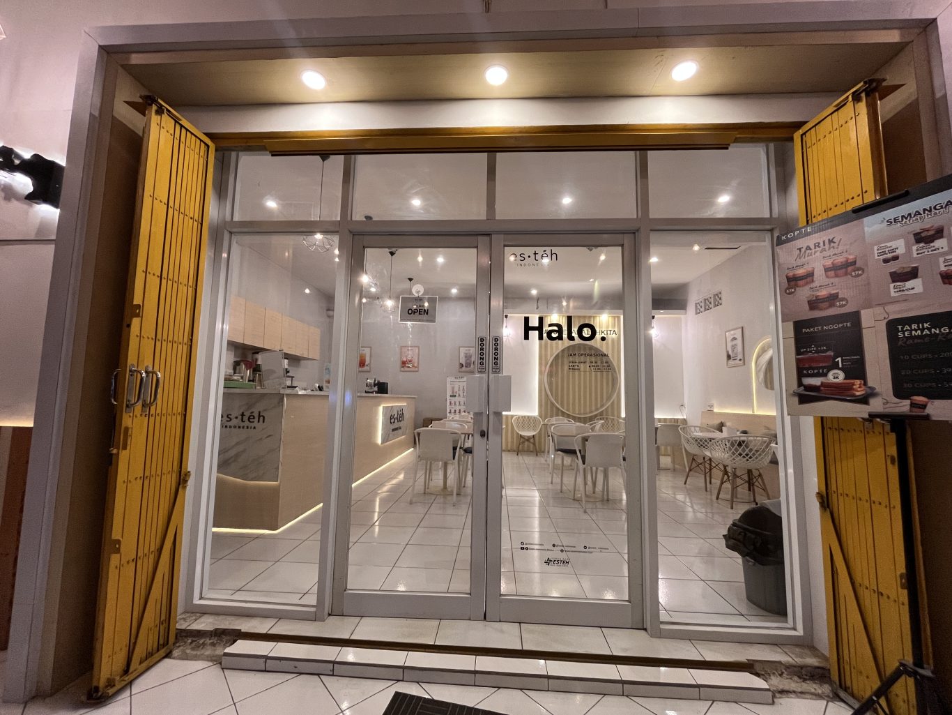The brightly lit interior of a modern Es Teh Indonesia shop in Waingapu, Sumba, is visible through its glass doors, flanked by partially open yellow security gates.