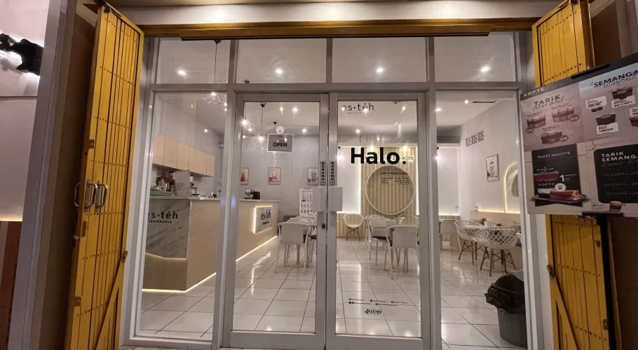 The brightly lit interior of a modern Es Teh Indonesia shop in Waingapu, Sumba, is visible through its glass doors, flanked by partially open yellow security gates.