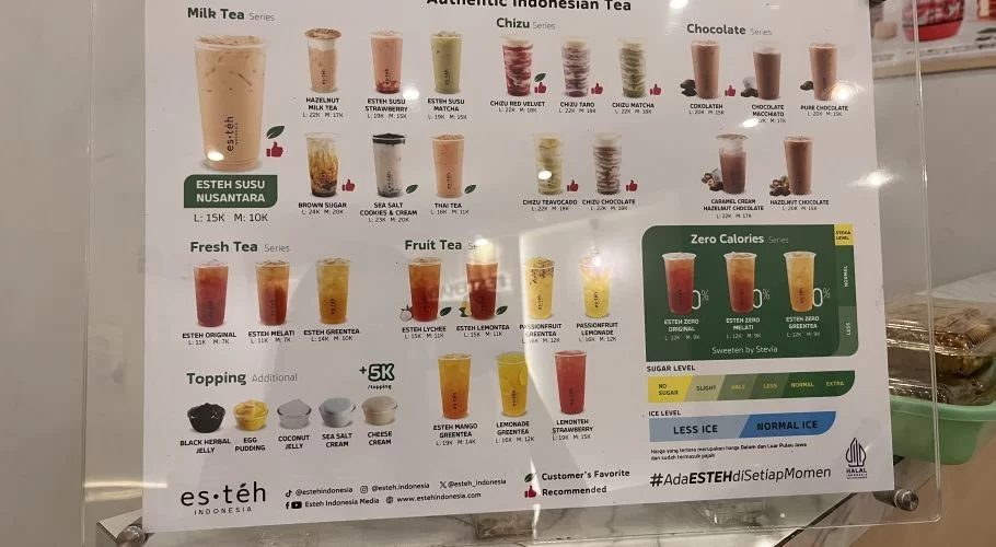 A brightly lit menu board for Es Teh Indonesia cafe displays numerous colorful tea beverages, prices, and customization options like toppings, sugar, and ice levels.