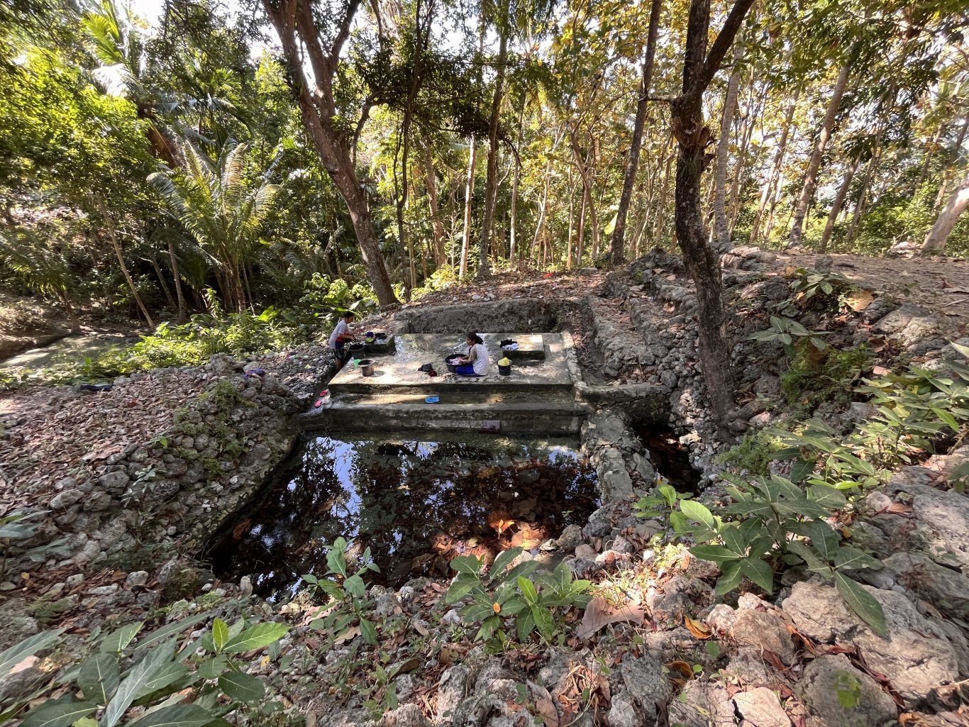 Two women are washing clothes at a multi-tiered concrete spring basin surrounded by dense green trees and rocky terrain under bright sunlight.