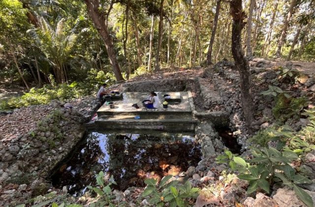 Two women are washing clothes at a multi-tiered concrete spring basin surrounded by dense green trees and rocky terrain under bright sunlight.