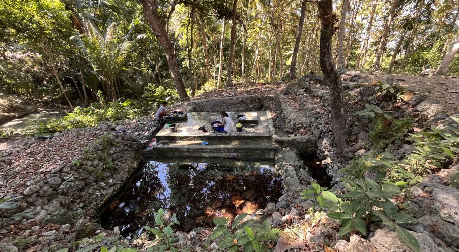 Two women are washing clothes at a multi-tiered concrete spring basin surrounded by dense green trees and rocky terrain under bright sunlight.