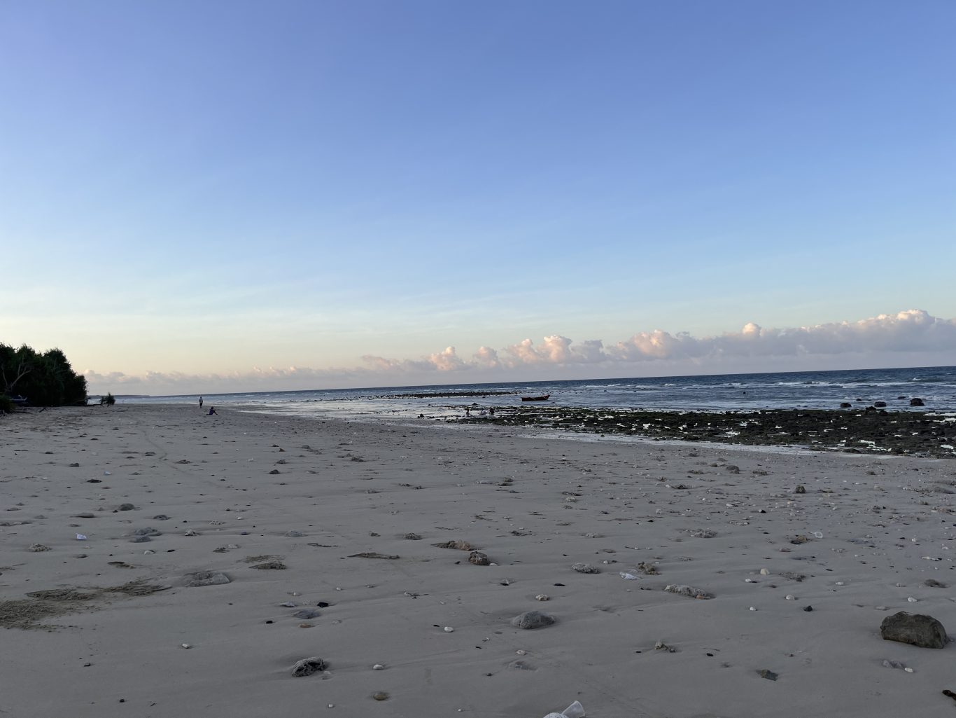 A wide, light-sandy beach stretches towards a calm blue ocean with a small boat, bordered by dark green trees and a clear sky with scattered clouds.