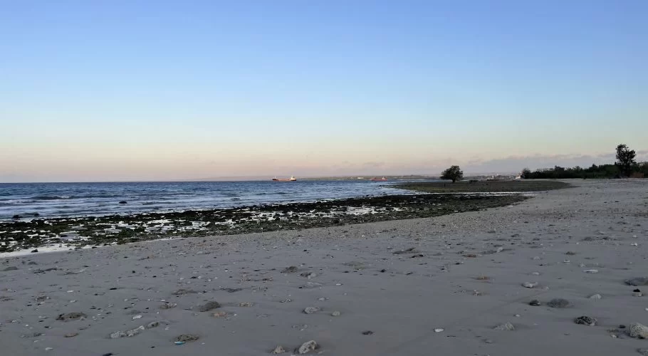 A wide view of Temu Beach at dusk, showing a rocky shoreline meeting light grey sand, gentle waves, and distant ships under a pale blue and orange sky.