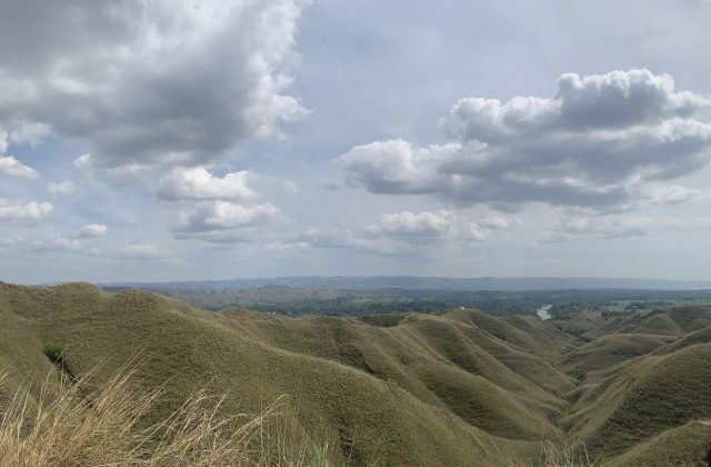 Vast rolling hills covered in dry, light brown grass stretch under a cloudy sky, with a distant valley and river visible.