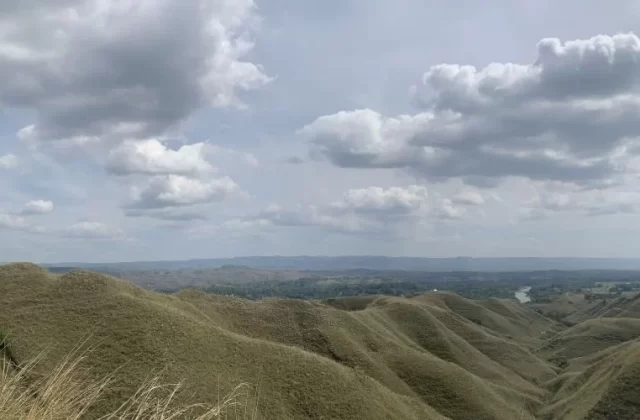 Vast rolling hills covered in dry, light brown grass stretch under a cloudy sky, with a distant valley and river visible.