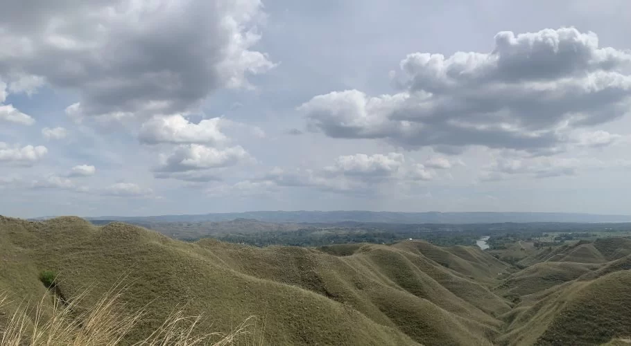 Vast rolling hills covered in dry, light brown grass stretch under a cloudy sky, with a distant valley and river visible.