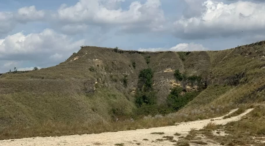 Dry, undulating brown hills with patches of green vegetation are visible under a partly cloudy sky, with a light dirt path in the foreground.