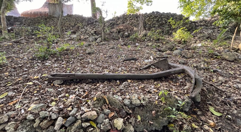 A centuries-old, dark metal anchor lies on dry, rocky ground covered in brown leaves, with a stone wall and trees behind it.