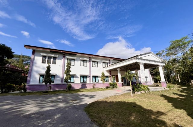 A two-story white building with purple trim and a columned entrance stands under a blue sky, surrounded by green trees and grass.
