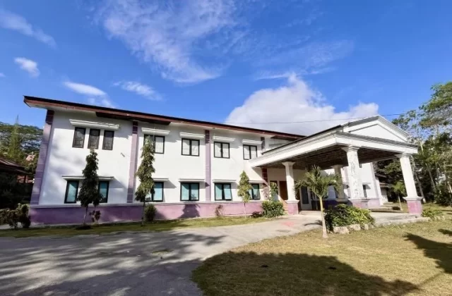 A two-story white building with purple trim and a columned entrance stands under a blue sky, surrounded by green trees and grass.