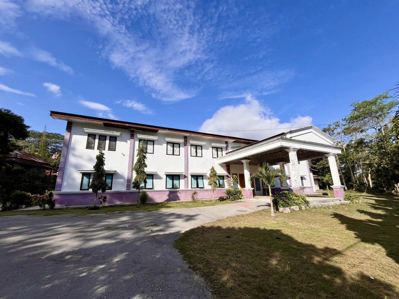 A two-story white building with purple trim and a columned entrance stands under a blue sky, surrounded by green trees and grass.
