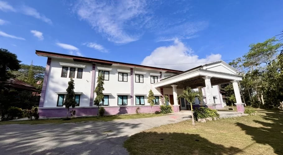 A two-story white building with purple trim and a columned entrance stands under a blue sky, surrounded by green trees and grass.