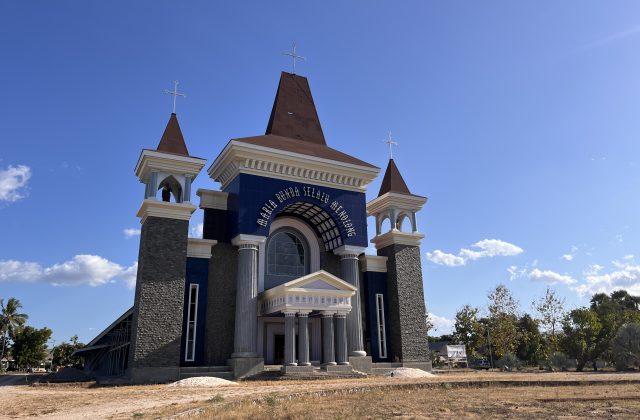 A large, modern Catholic church with a dark blue facade, white trim, and three cross-topped towers stands under a clear blue sky in Waingapu, Sumba.