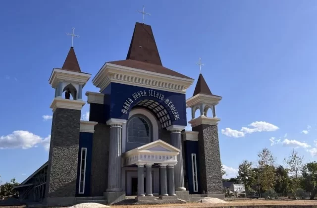 A large, modern Catholic church with a dark blue facade, white trim, and three cross-topped towers stands under a clear blue sky in Waingapu, Sumba.