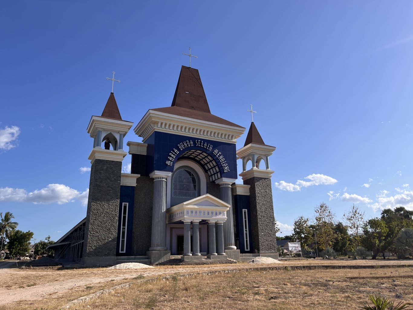 A large, modern Catholic church with a dark blue facade, white trim, and three cross-topped towers stands under a clear blue sky in Waingapu, Sumba.