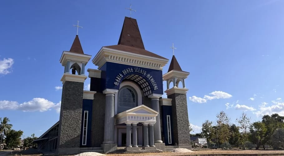 A large, modern Catholic church with a dark blue facade, white trim, and three cross-topped towers stands under a clear blue sky in Waingapu, Sumba.