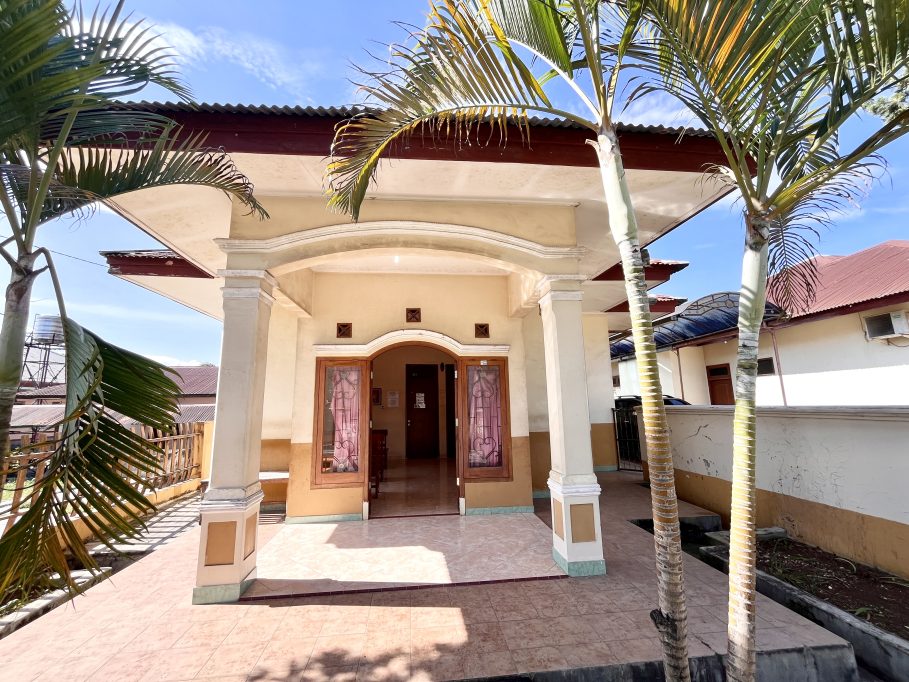 The Lende Moripa Doctor Clinic in Waikabubak features a cream-colored facade with an arched entrance, flanked by two palm trees under a blue sky.