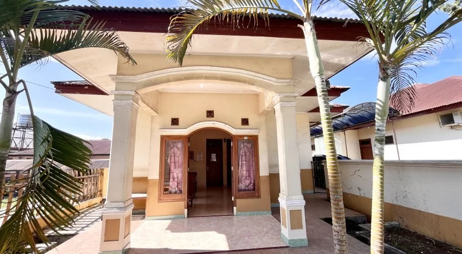 The Lende Moripa Doctor Clinic in Waikabubak features a cream-colored facade with an arched entrance, flanked by two palm trees under a blue sky.