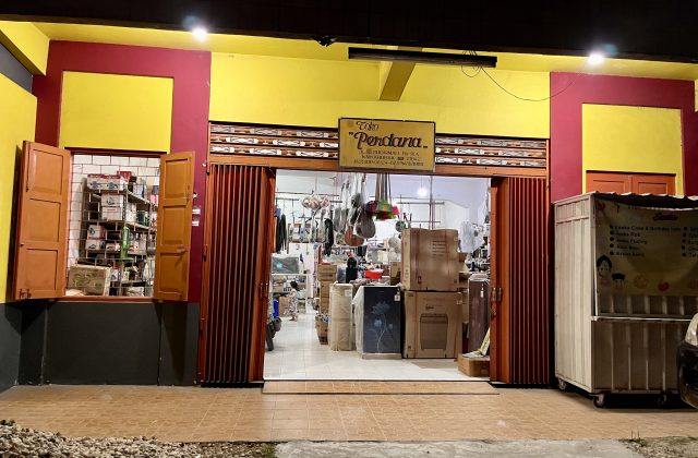 A brightly lit storefront at night, featuring a yellow and red facade with "Toko Perdana" sign. The open entrance reveals an interior stocked with household goods and a food cart outside.