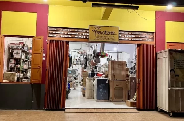 A brightly lit storefront at night, featuring a yellow and red facade with "Toko Perdana" sign. The open entrance reveals an interior stocked with household goods and a food cart outside.