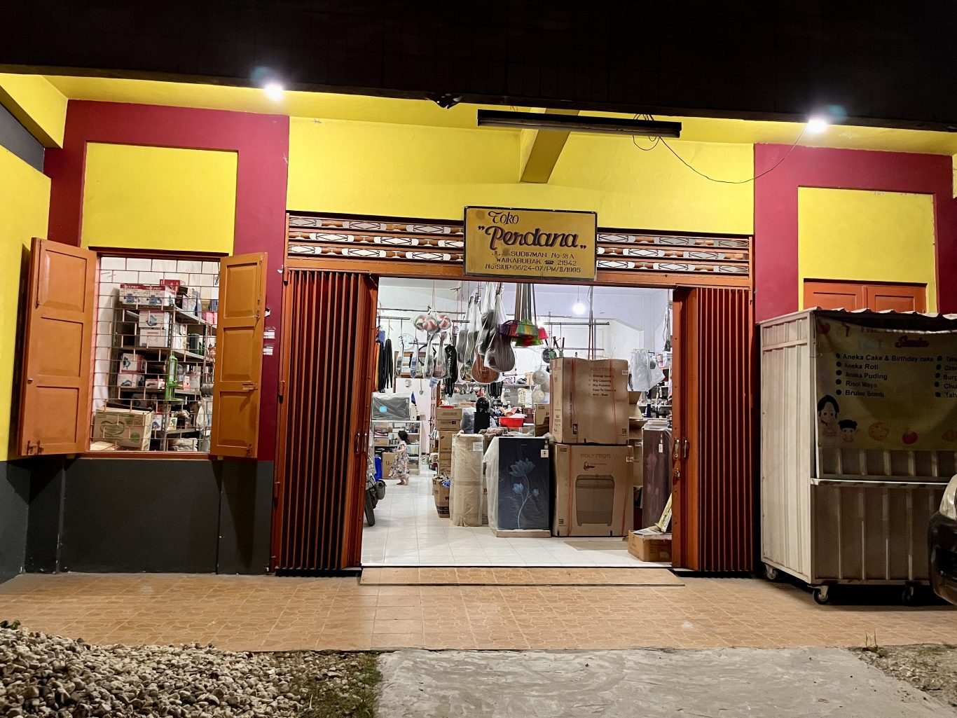 A brightly lit storefront at night, featuring a yellow and red facade with "Toko Perdana" sign. The open entrance reveals an interior stocked with household goods and a food cart outside.