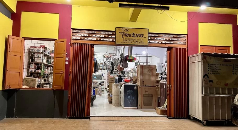 A brightly lit storefront at night, featuring a yellow and red facade with "Toko Perdana" sign. The open entrance reveals an interior stocked with household goods and a food cart outside.