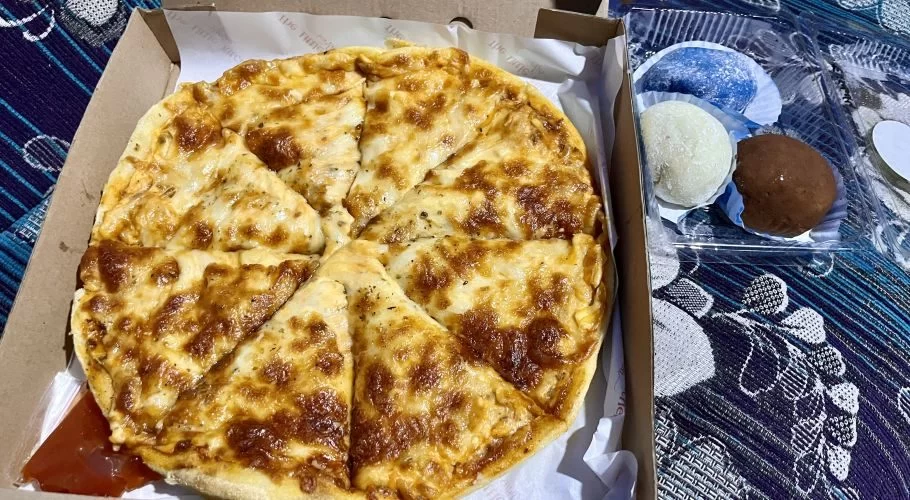 A golden-brown cheesy pizza, sliced in a cardboard box, sits next to a container of blue, white, and brown mochi on a patterned blue fabric.