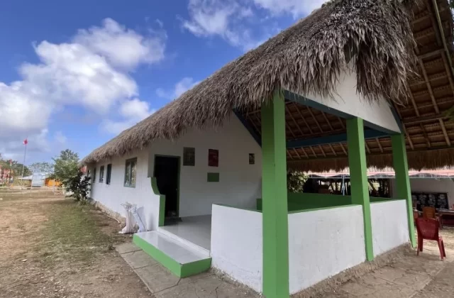 A white clinic building with a traditional thatched roof and green accents stands under a blue sky with clouds, surrounded by dry ground and Indonesian flags.