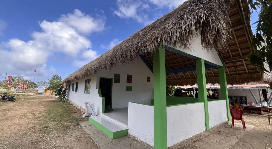 A white clinic building with a traditional thatched roof and green accents stands under a blue sky with clouds, surrounded by dry ground and Indonesian flags.