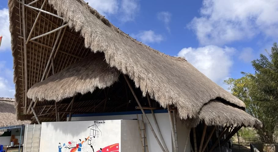 A building with a large, multi-layered thatched roof and exposed bamboo structure stands under a blue sky, featuring a colorful mural on its white wall.