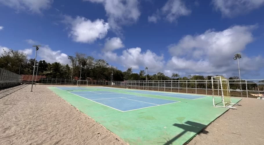A vibrant blue and green multi-sport court is surrounded by sandy ground, a white fence, and green trees under a bright blue sky with white clouds.