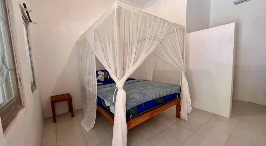 A simple homestay bedroom features a wooden bed with a blue blanket and white mosquito net, a small stool, and an air conditioning unit.