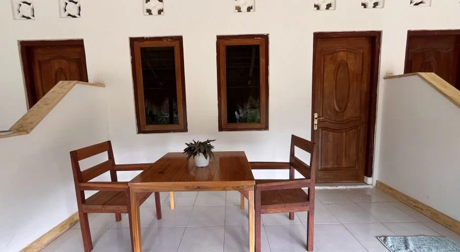 A wooden table with two chairs sits on a tiled patio outside a white building with several dark wooden doors and windows, and decorative wall vents.